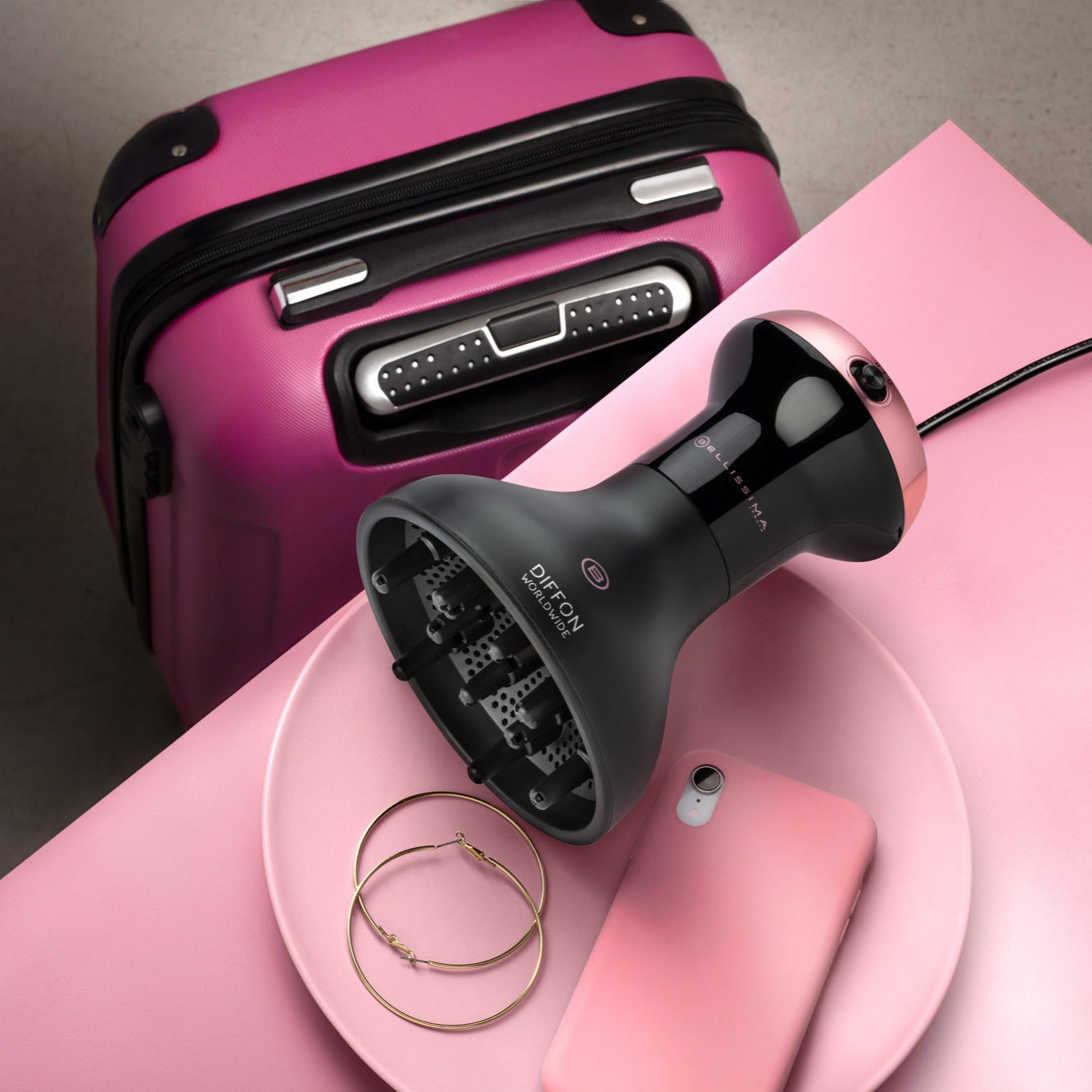 Black Diffon Worldwide curly hair dryer on pink surface with pink suitcase in background